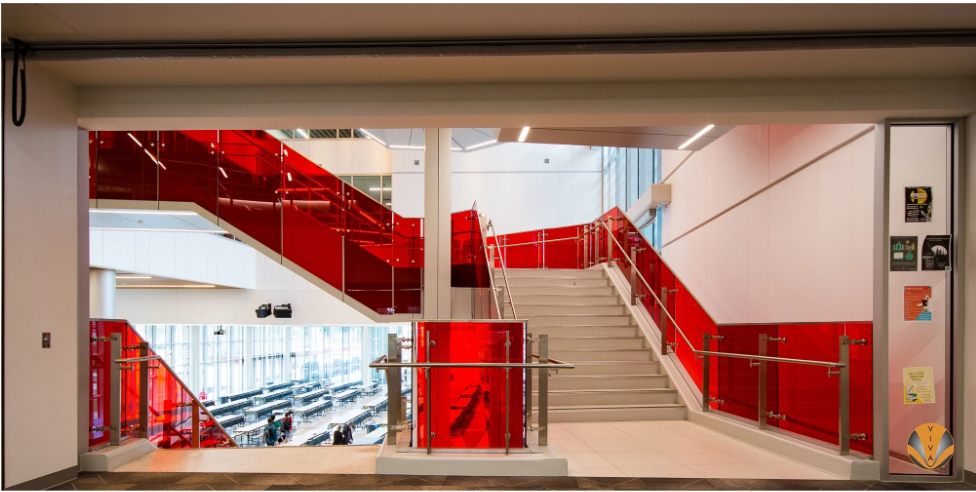 Modern school interior with bold red glass stair railings and stainless steel handrails overlooking a cafeteria.
